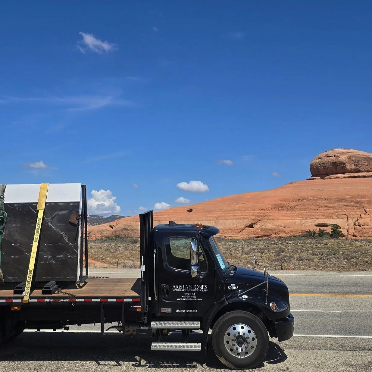 A black flatbed truck with embedded logos and text reading 'Arista Stones' parked on a road with a desert landscape and red rock formations in the background under a bright blue sky with a few white clouds.