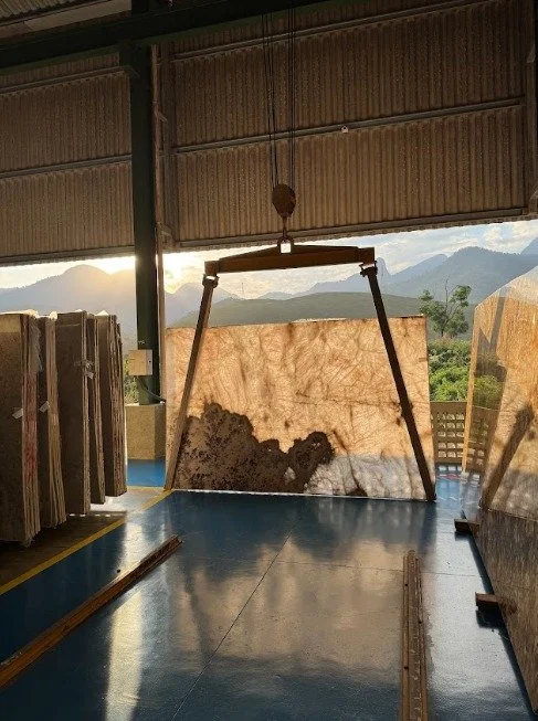 Large golden stone slab hangs from lifting equipment inside a warehouse, with sunlight and mountains visible beyond.