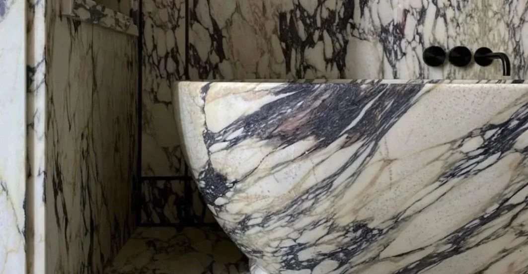 White marble vessel sink with bold black and gray veining against matching marble walls and a black wall-mounted faucet.