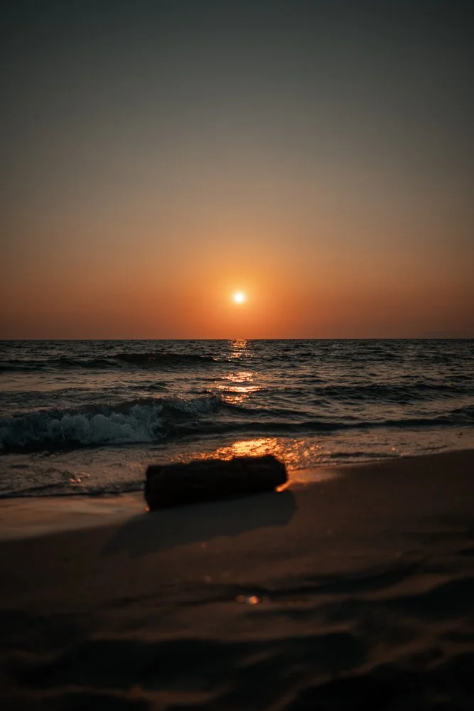 Sonnenuntergang über dem Meer mit Wellen und einem Stein im Vordergrund am Strand.