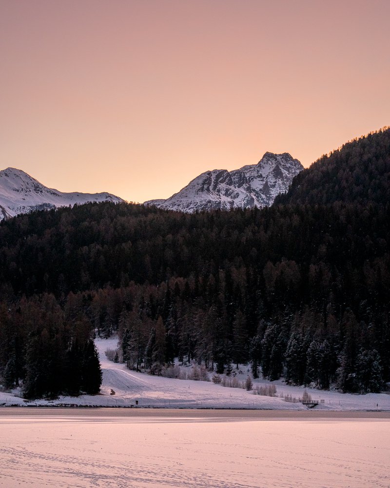 Berglandschaft mit schneebedeckten Bergen, bewaldeter Hügel, gefrorenem See und Sonnenuntergangshimmel.