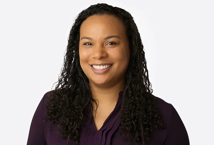 A woman with long curly dark hair smiling, wearing a dark purple blouse, against a plain light background.