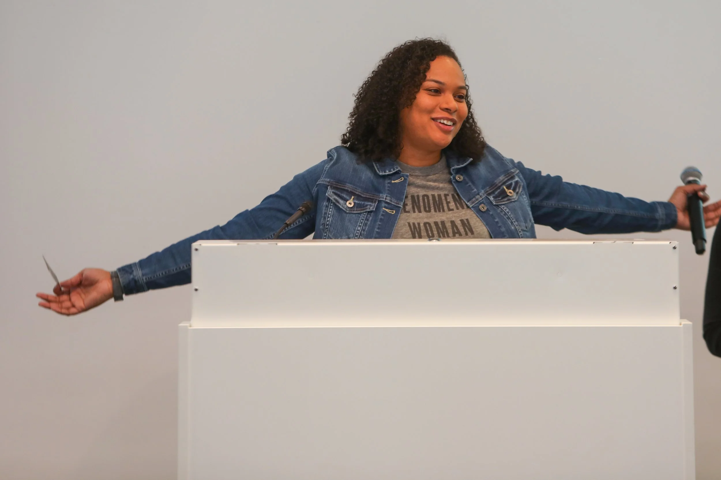 A woman with curly hair wearing a denim jacket and a gray t-shirt with the words 'Feminism Woman' stands behind a white podium, smiling with her arms extended to the sides, holding a microphone in her right hand.