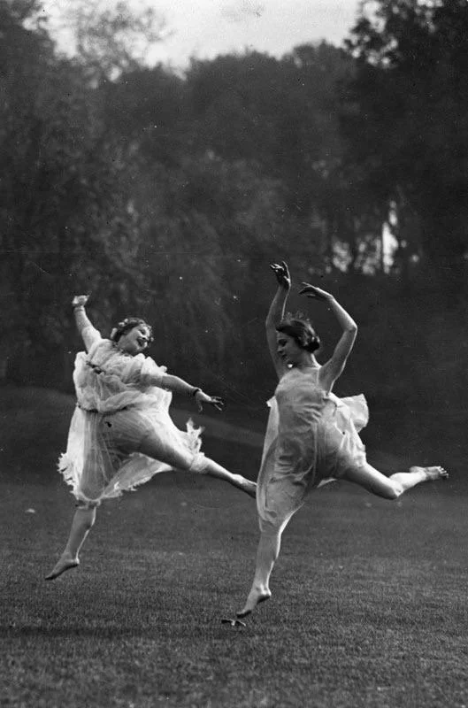 Two women in flowing dresses dance on a grassy field outdoors, captured mid-movement with arms raised and legs extended.