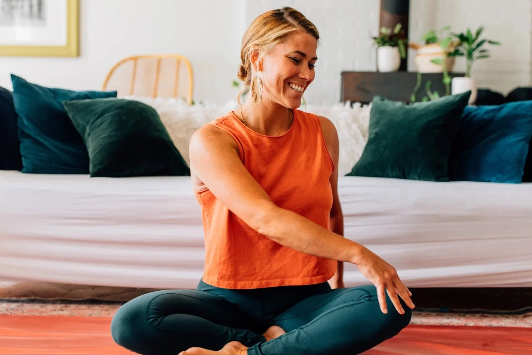 A woman with blonde hair, wearing an orange sleeveless top and blue yoga pants, sits cross-legged on a yoga mat in a living room, smiling while stretching her arm.