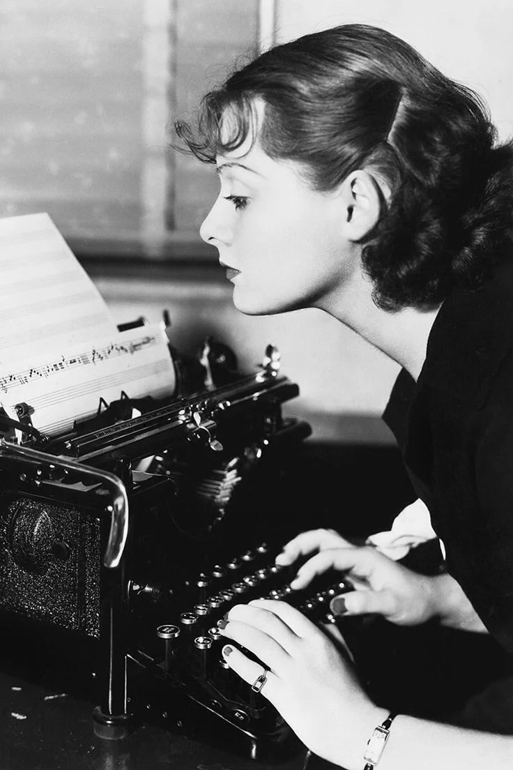 A woman with wavy, shoulder-length hair typing on a vintage typewriter with sheet music.