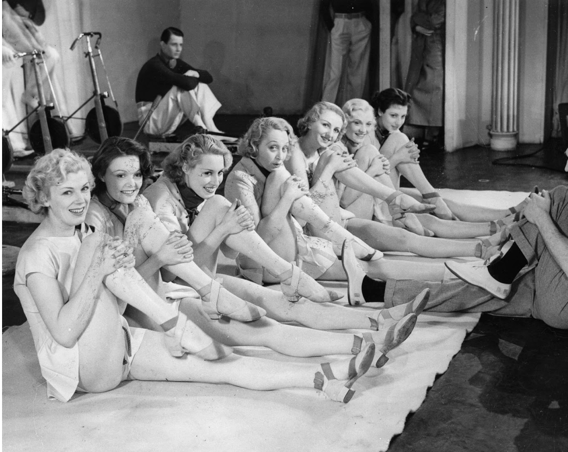 A group of seven young women in dance costumes sitting in a line on the floor, holding one leg with their arms. There are a few men in the background, with one sitting on a bench and others standing.