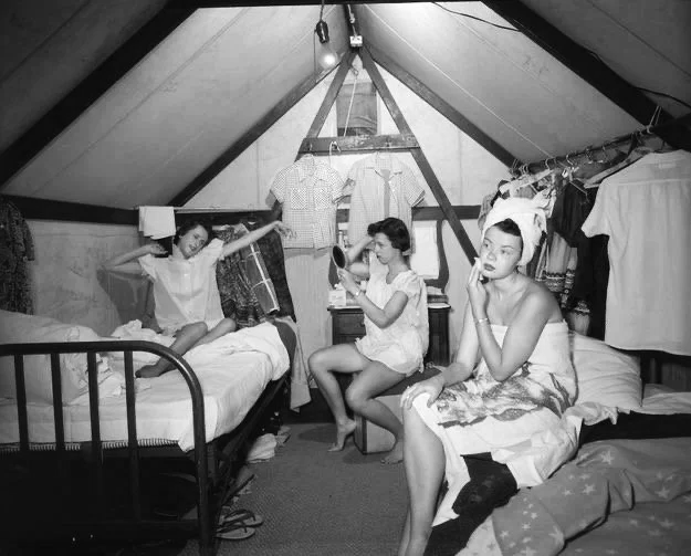 Three women in a cozy attic bedroom with clothes hanging on a rack and a small side table, one sitting on the bed using a hairdryer, one sitting on the bed cleaning her face, and one sitting on another bed talking on the phone.