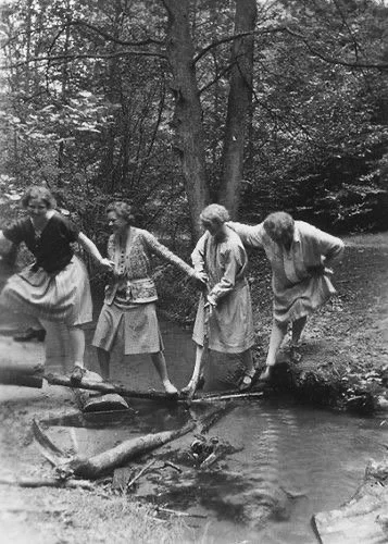 Four girls holding hands crossing a small wooden bridge over a creek in a wooded area, in a black and white photo.