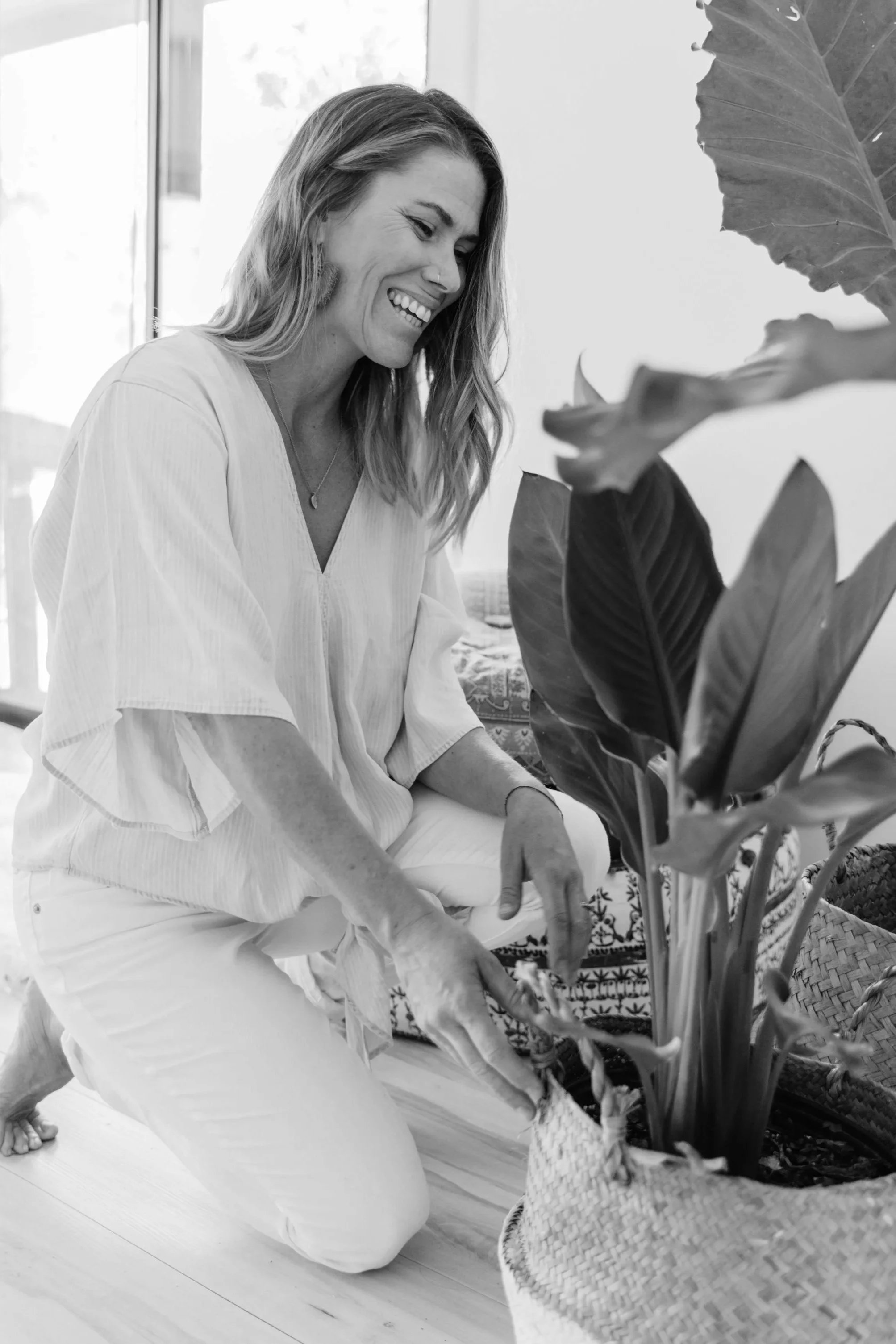 A woman with shoulder-length hair smiling and wearing a nose ring, sitting on the floor, interacting with large potted plants with broad leaves in a well-lit indoor space.