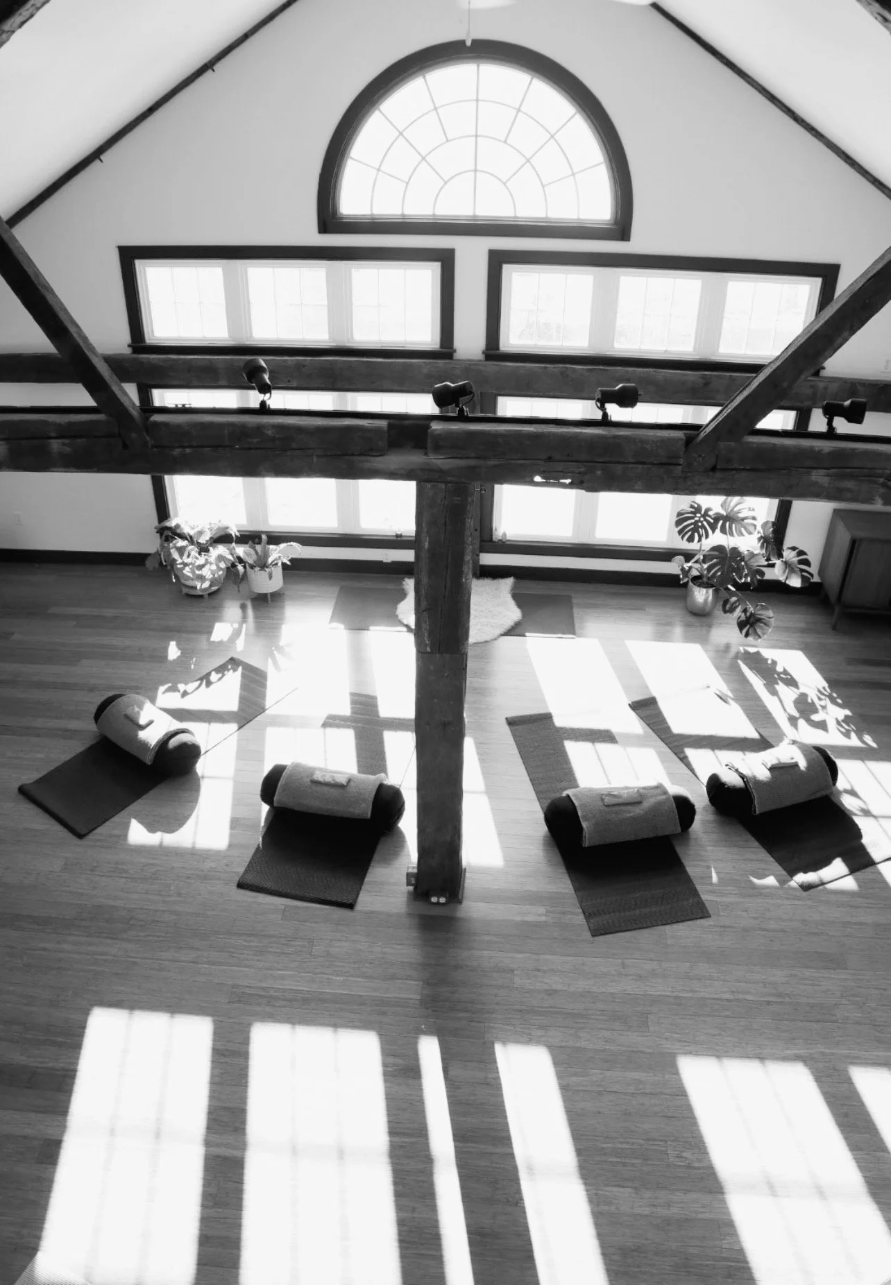A cozy yoga or meditation space with yoga mats and rolled-up blankets arranged in a circle. Sunlight streams through windows, casting shadows on the wooden floor, with plants and a small table in the background.