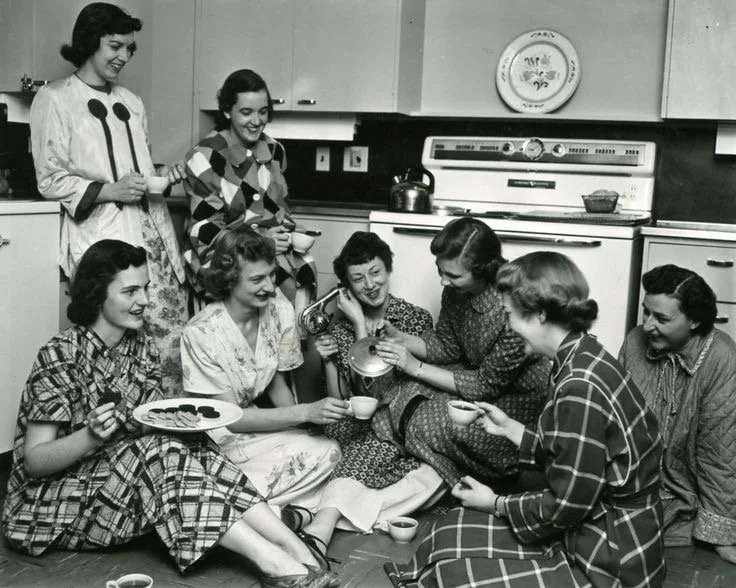 Group of women in vintage clothing having tea and cookies in a kitchen.