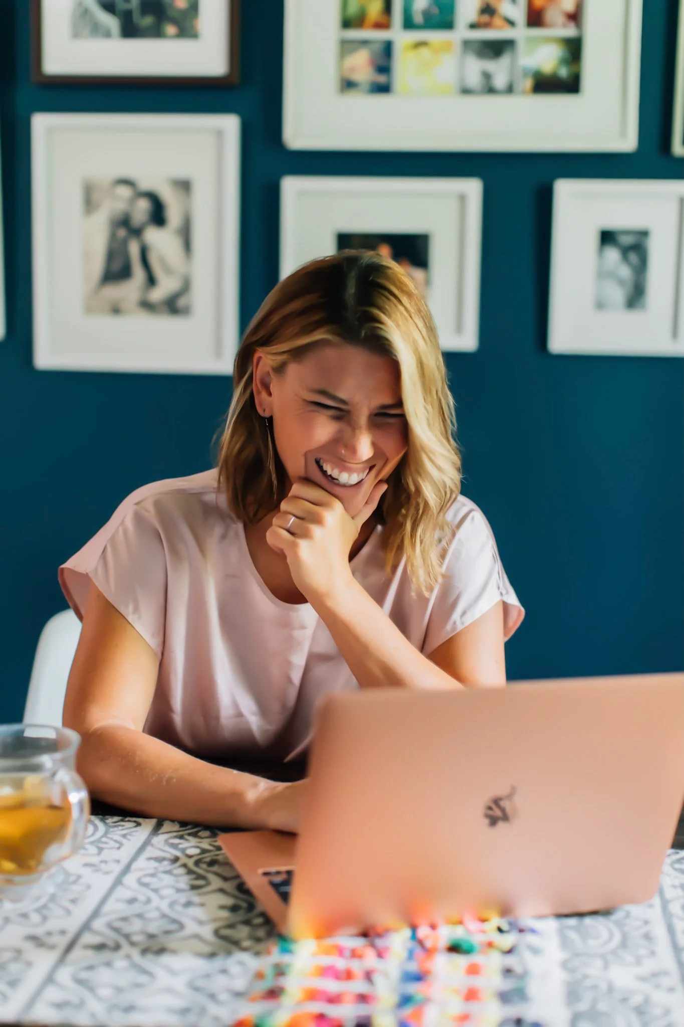 A woman with blonde hair is sitting at a table, smiling and laughing, while looking at a laptop. There are framed photos on a blue wall behind her, and a colorful spread of objects on the table.