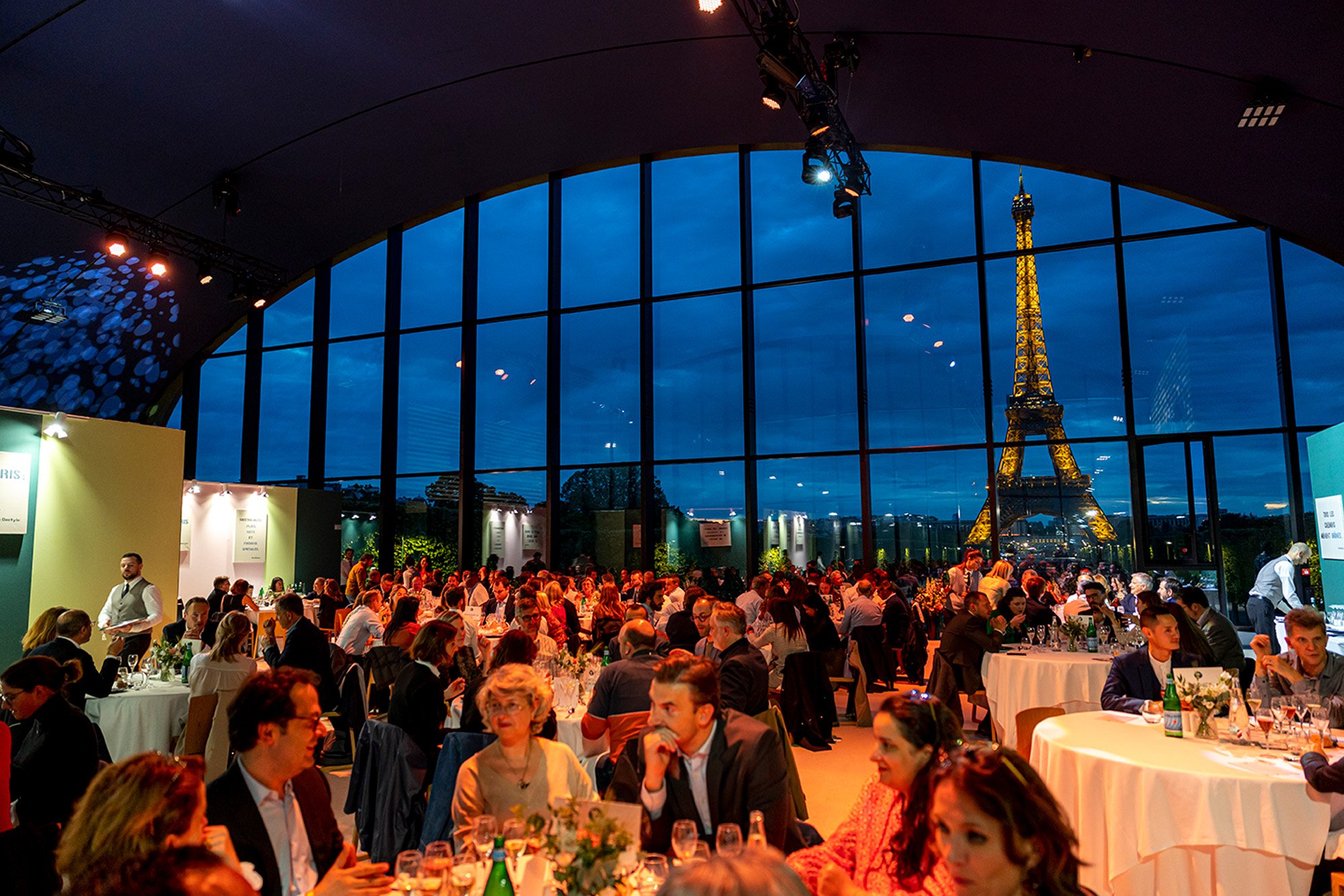 Un grand événement ou dîner dans une salle avec de grandes fenêtres donnant sur la Tour Eiffel illuminée de nuit à Paris, avec de nombreuses personnes assises à des tables.