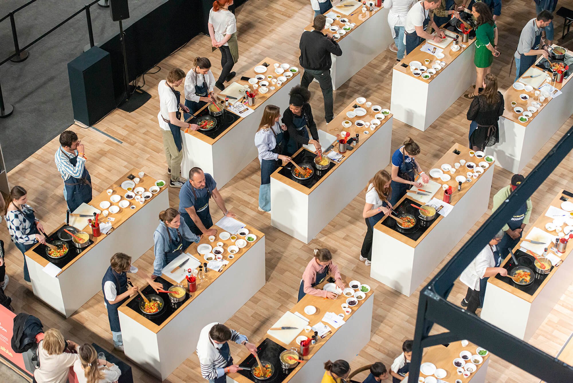 Cours de cuisine avec plusieurs personnes préparant des plats sur des îlots dans une grande salle.