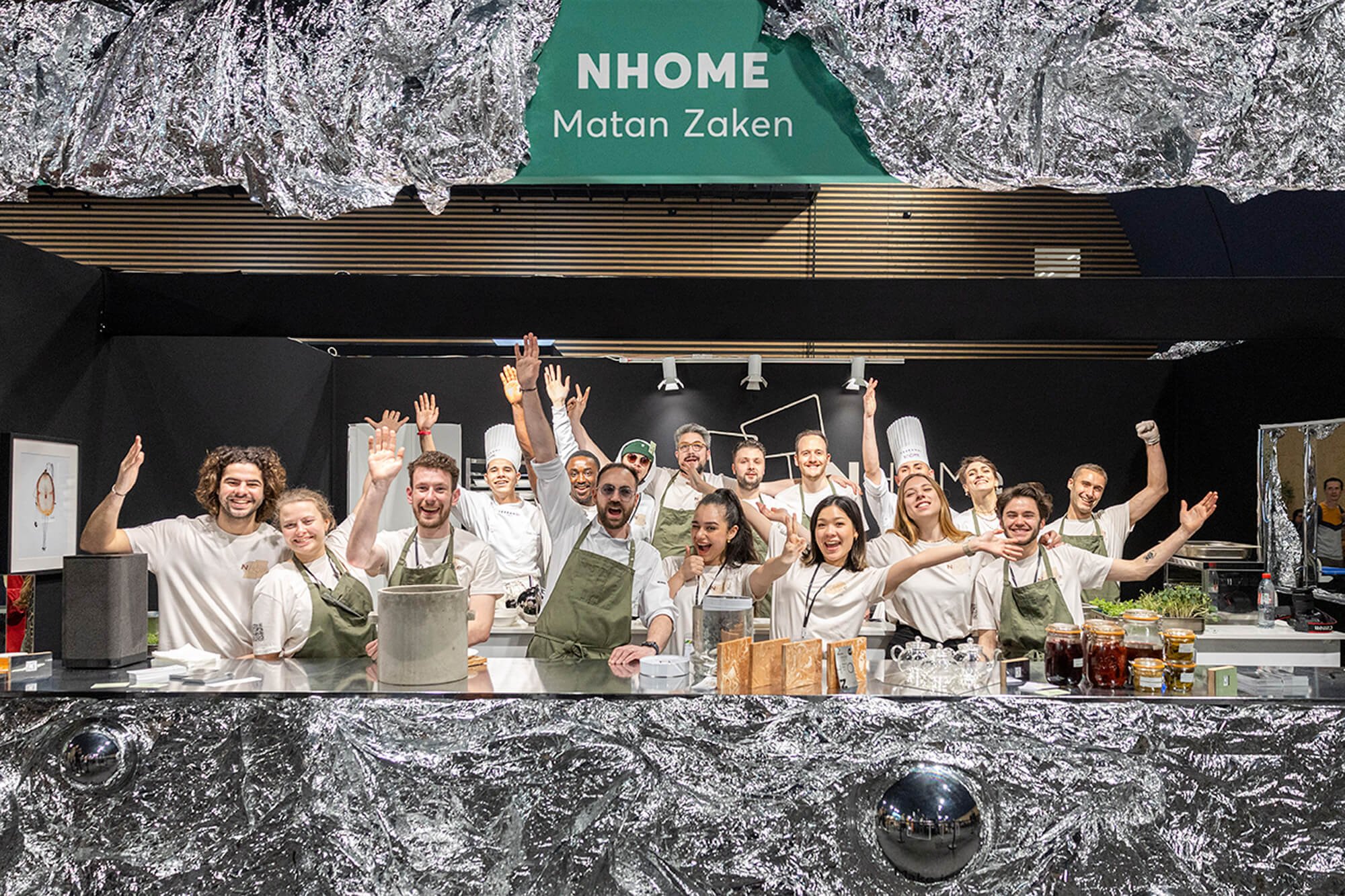 Groupe de cuisiniers souriants et levant les bras dans une cuisine professionnelle avec décor en aluminium et pancarte 'NHOME Matan Zaken' suspendue au plafond.