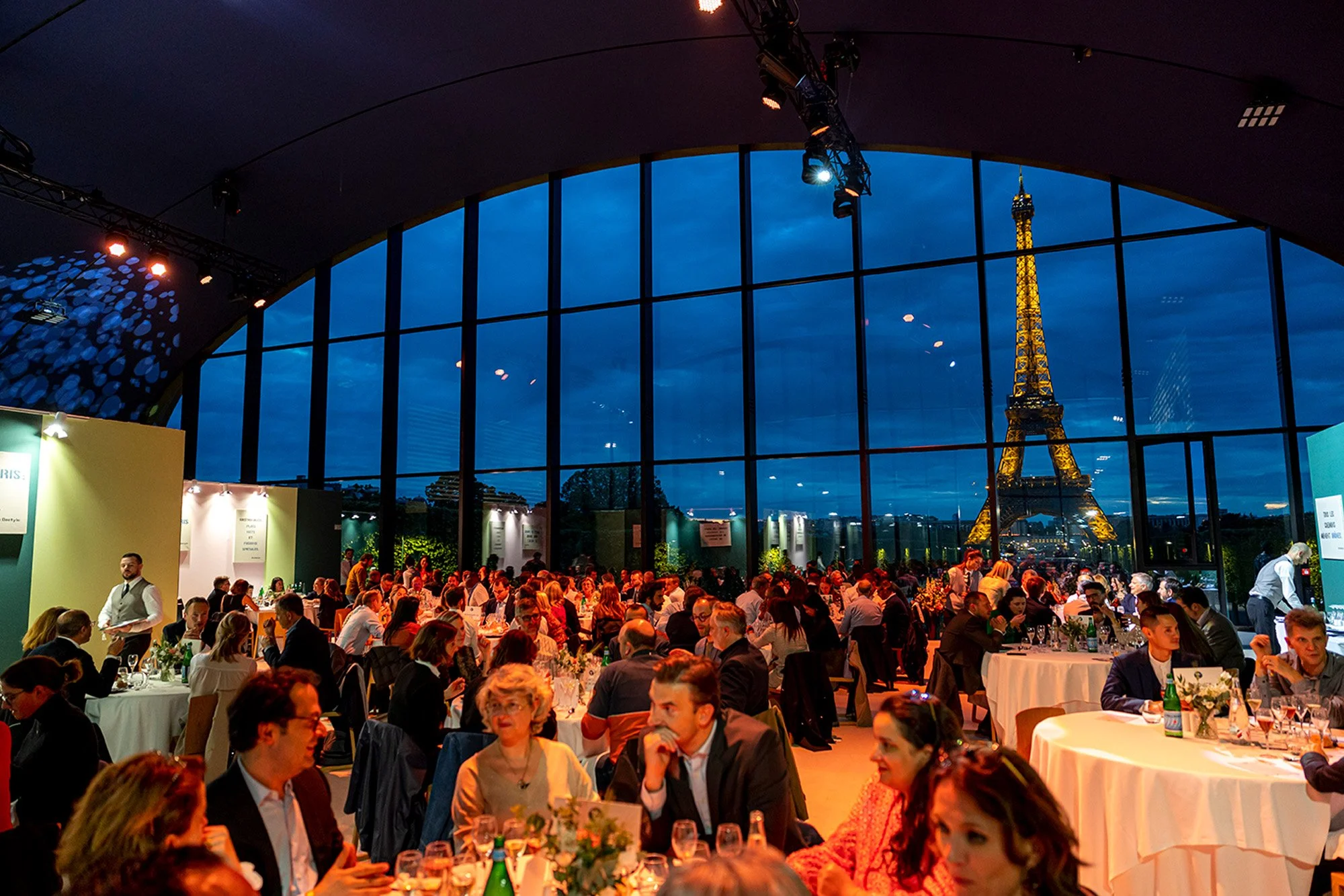 Grande salle de banquet avec vue sur la Tour Eiffel illuminée à Paris, remplie de nombreuses personnes assises à des tables rondes lors d'un événement ou dîner en soirée.