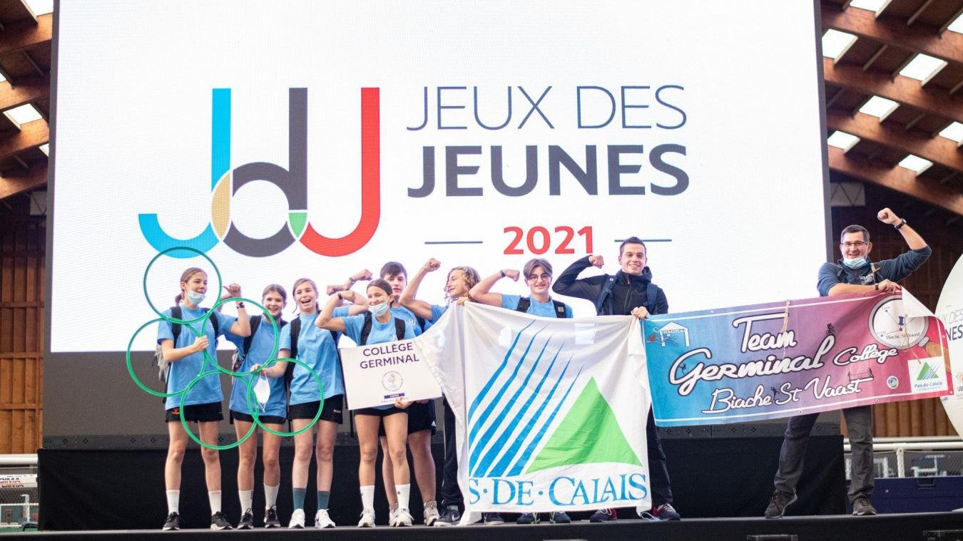 Groupe de jeunes filles et deux jeunes hommes sur scène en train de célébrer leur victoire lors des Jeux des Jeunes 2021, tenant des drapeaux et portant des médailles, sous une grande bannière