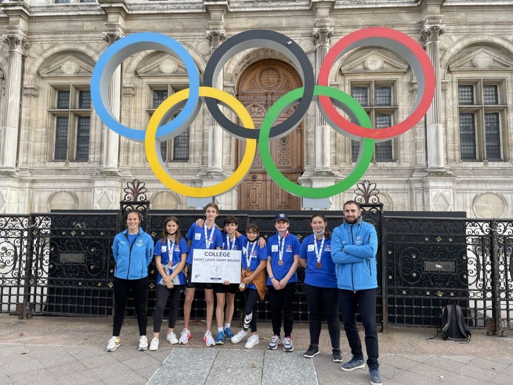 Groupe de jeunes athlètes posant sous les anneaux olympiques devant un bâtiment historique.