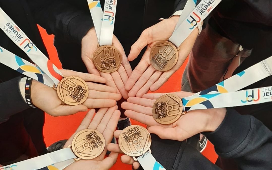 Groupe de six mains tenant des médailles de bronze pour les Jeux Jeunes 2022, sur fond rouge.