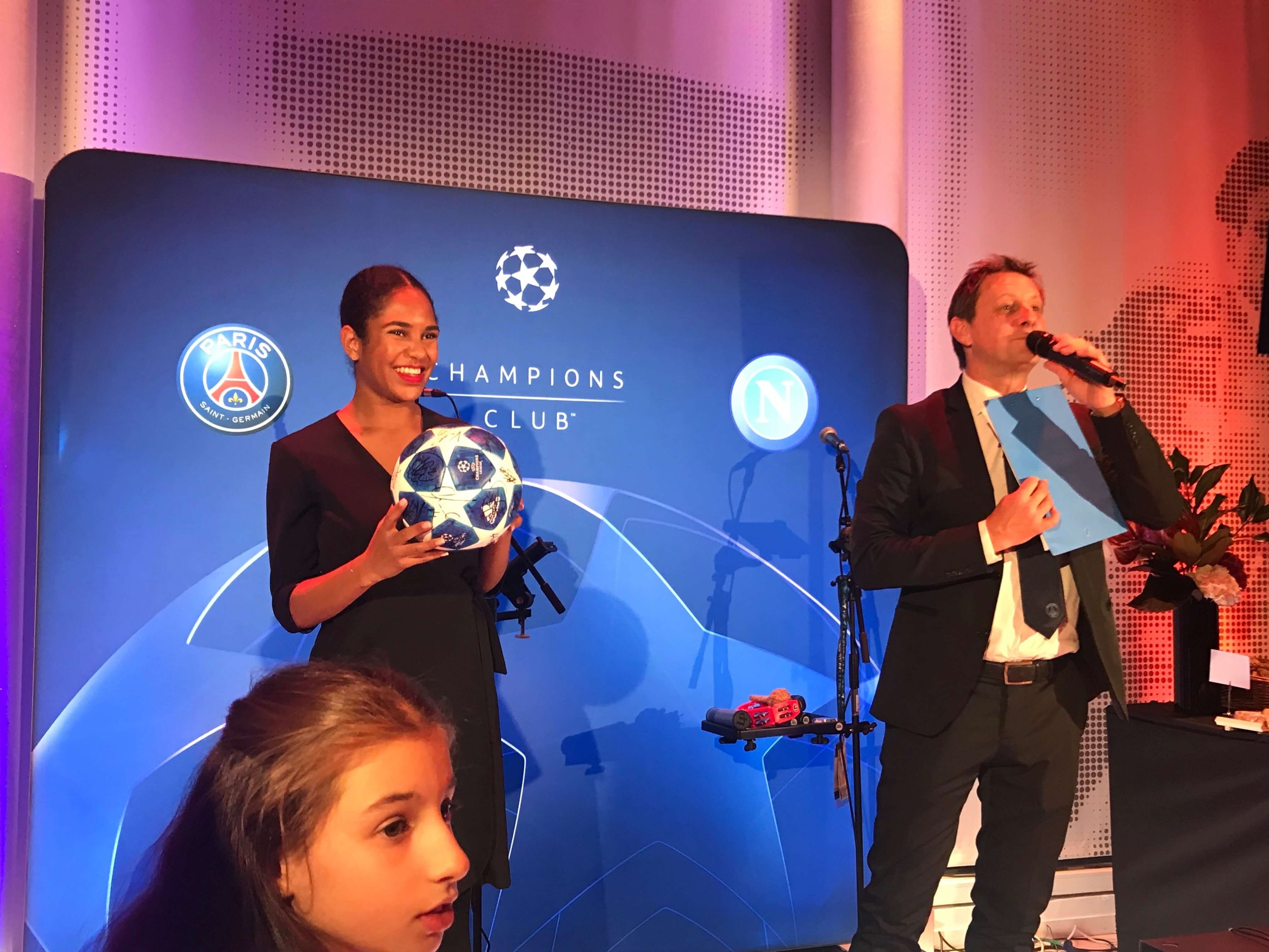 Une femme souriante tenant un ballon de football de la Ligue des champions à une conférence de presse, avec un homme parlant dans un micro et deux logos de football sur un fond bleu.