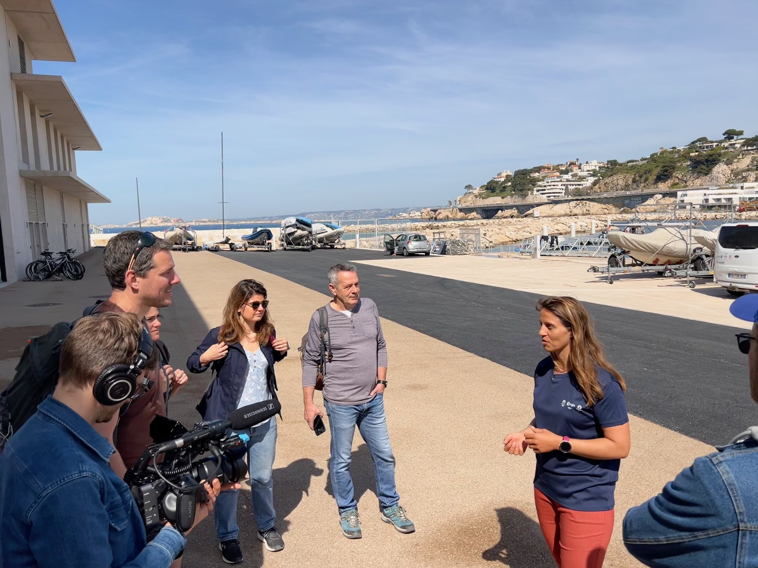 Groupe de personnes en conversation près d'un port avec des bateaux et des voitures en arrière-plan, en plein jour avec ciel clair.