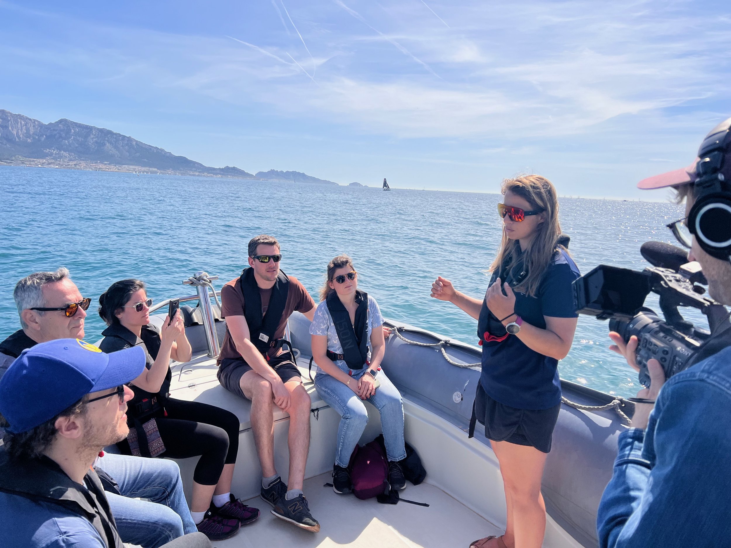 Groupe de personnes assises sur un bateau, une femme debout parle et filme une discussion, en arrière-plan la mer et des montagnes sous un ciel ensoleillé.