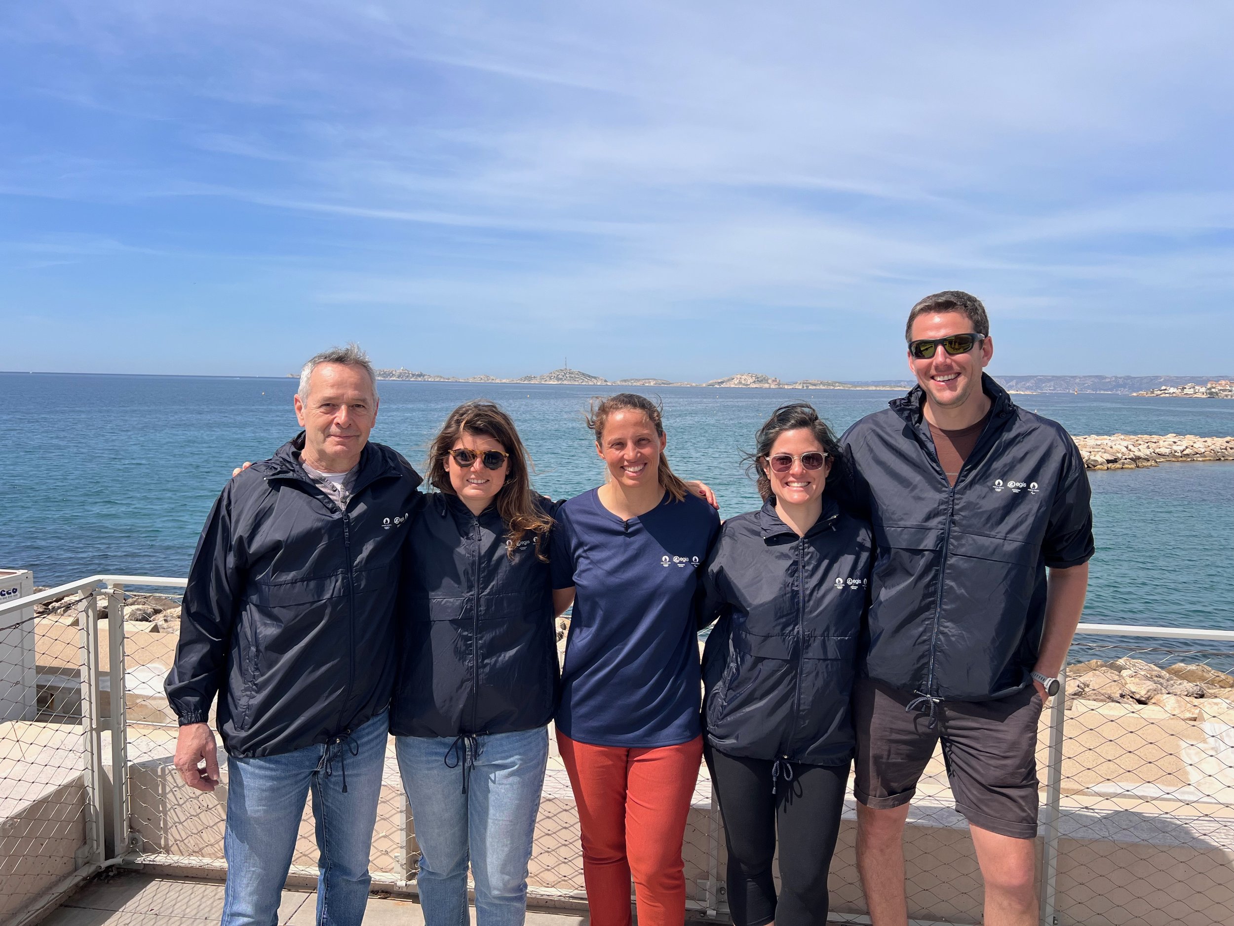 Groupe de cinq personnes souriantes posant près de la mer, portant des vestes noires, avec un ciel bleu et la mer en arrière-plan.