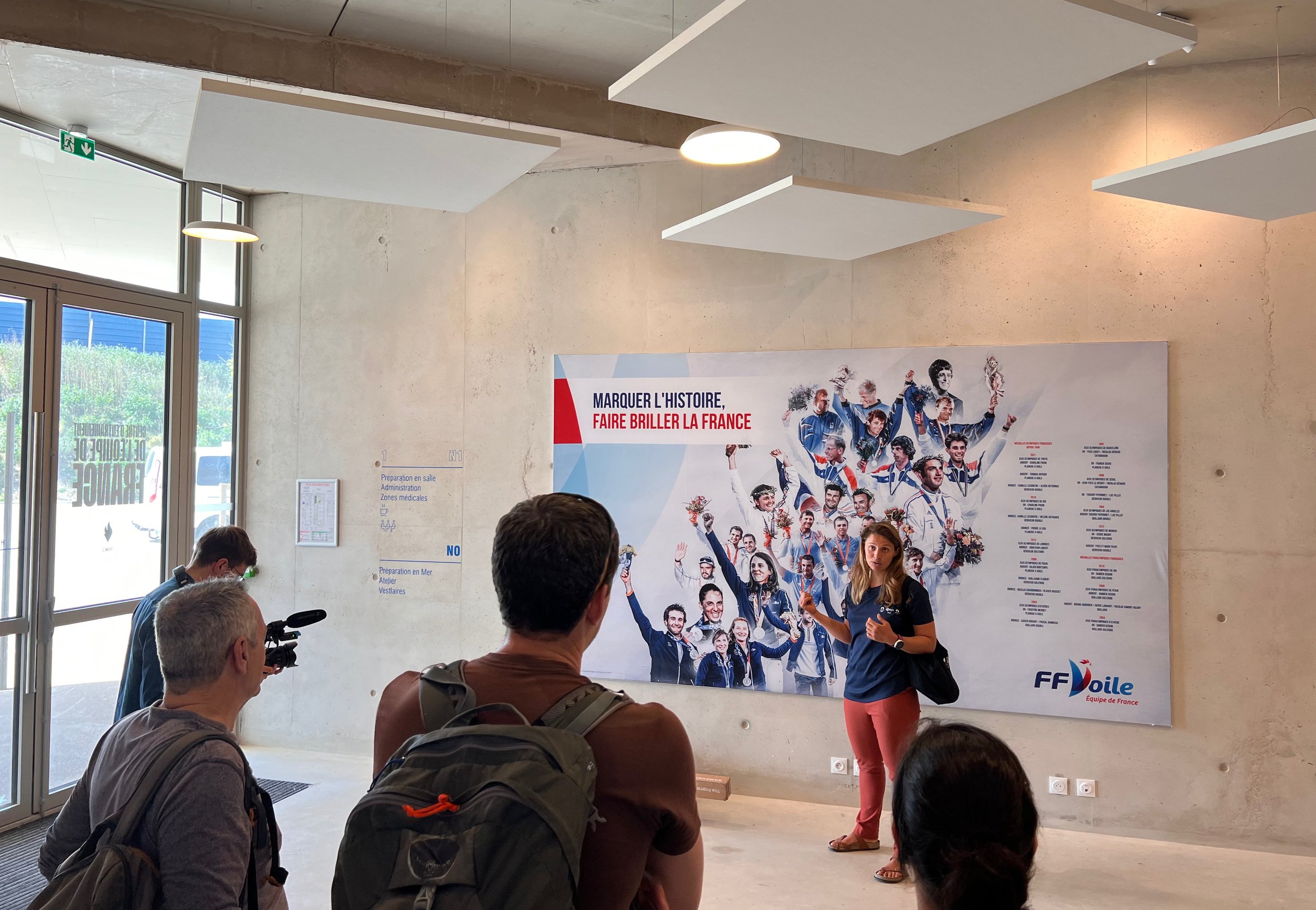 Une jeune femme explique quelque chose à un groupe de personnes dans une salle d'accueil, devant une affiche représentant une équipe de rugby française. Il y a des fenêtres à gauche, et le mur derrière la femme est en béton brut.