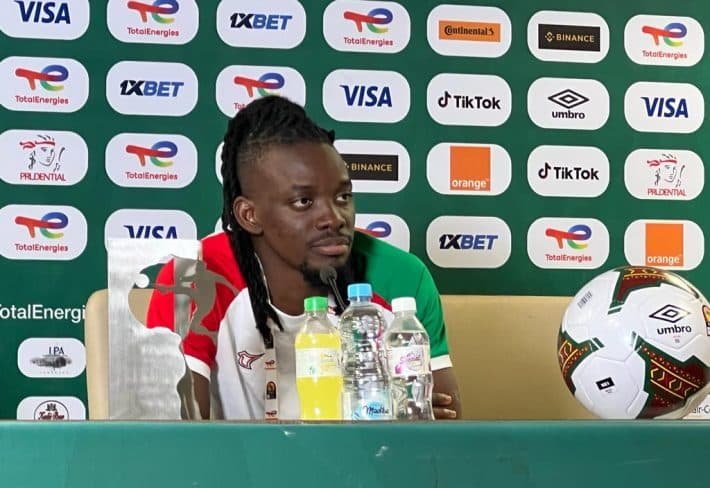 Un homme assis à une table lors d'une conférence de presse, avec plusieurs bouteilles d'eau et une balle de football devant lui, derrière un panneau publicitaire avec divers logos.