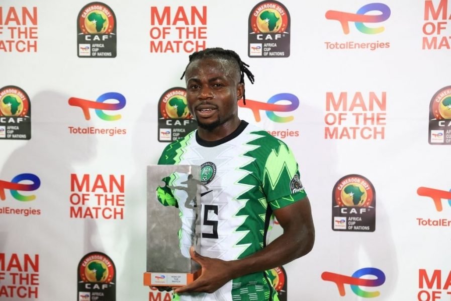 Un joueur de football camerounais en costume vert et blanc tenant un trophée lors d'une cérémonie, avec un fond affichant 'MAN OF THE MATCH' et les logos de la CAF et TotalEnergies.