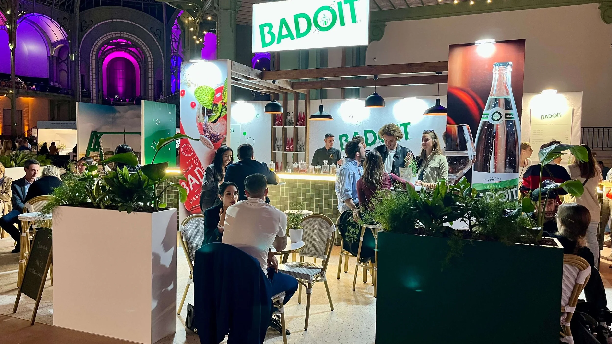 Vue de nuit du bar Badoit, crée par l'agence Agon à l'occasion de Taste of Paris 2025. On distingue des visiteurs attablés en train de déguster des cocktails sans alcool.