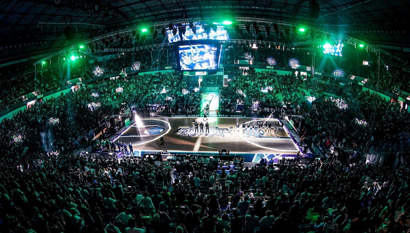 Une salle de basketball pleine avec un match en cours, lumières vertes et écrans géants qui diffusent la partie.