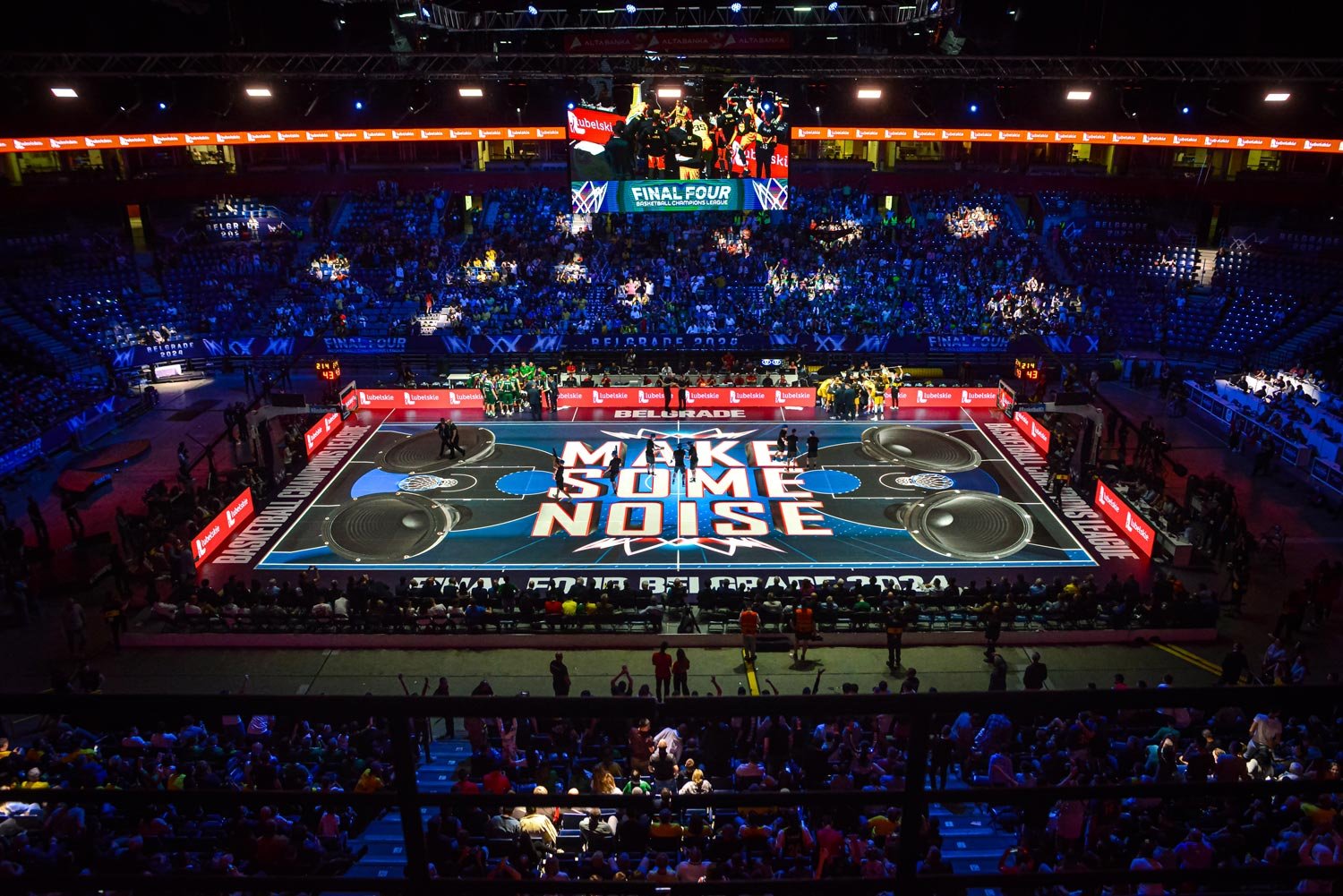 Un terrain de basketball transformé pour un événement, avec un écran géant affichant "Final Four" et des joueurs rassemblés, dans une salle remplie de spectateurs, et le message "Make Some Noise" peint sur le sol.