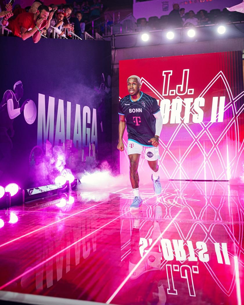 Un joueur de basketball portant un maillot bleu et blanc court sur un parquet brillant, avec un fond rouge moderne affichant 'J.J. Oatsis II'. Il y a des spectateurs au-dessus, et des lumières et fumée créent une ambiance dynamique.