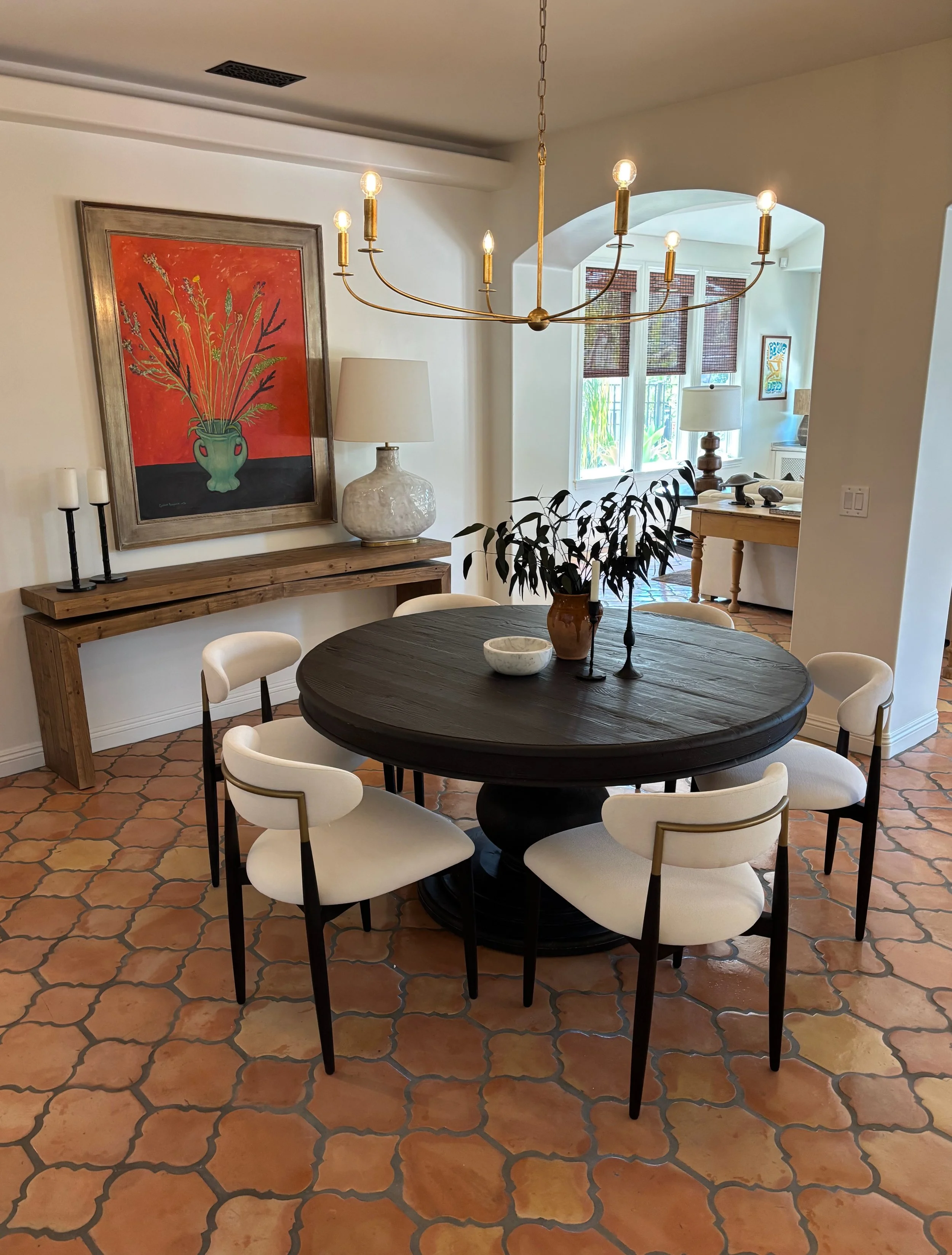A dining room with a round black table surrounded by six white upholstered chairs. A large chandelier hangs above the table. On the table, there is a vase with dark green foliage and two candlesticks. In the background, a wooden sideboard with a large lamp and artwork on the wall.