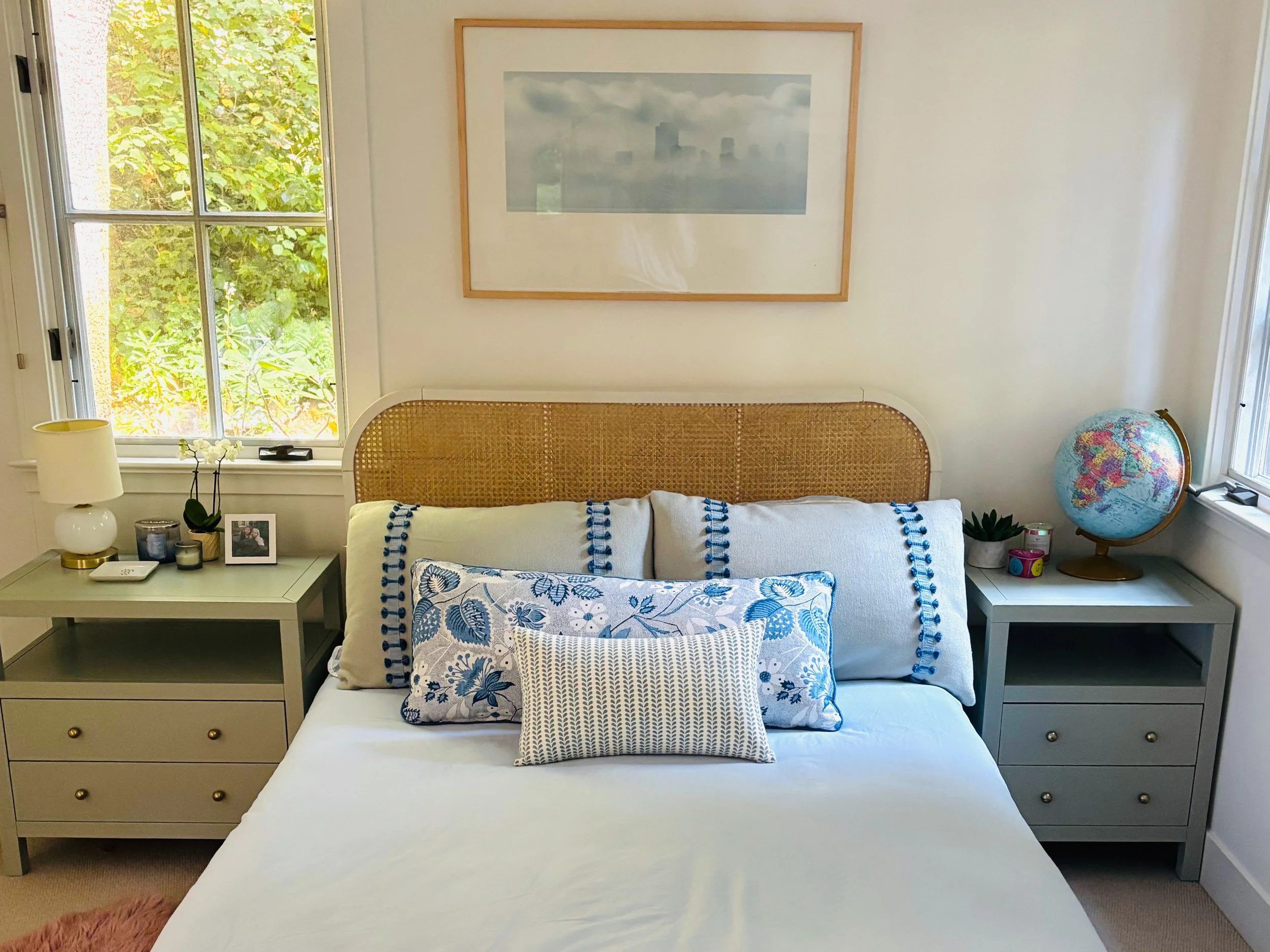 A neatly made bedroom with a bed featuring blue and white floral and striped pillows, white bedding, and a rattan headboard. Two light gray nightstands with drawers flank the bed; the left nightstand has a table lamp, a flower arrangement, and framed