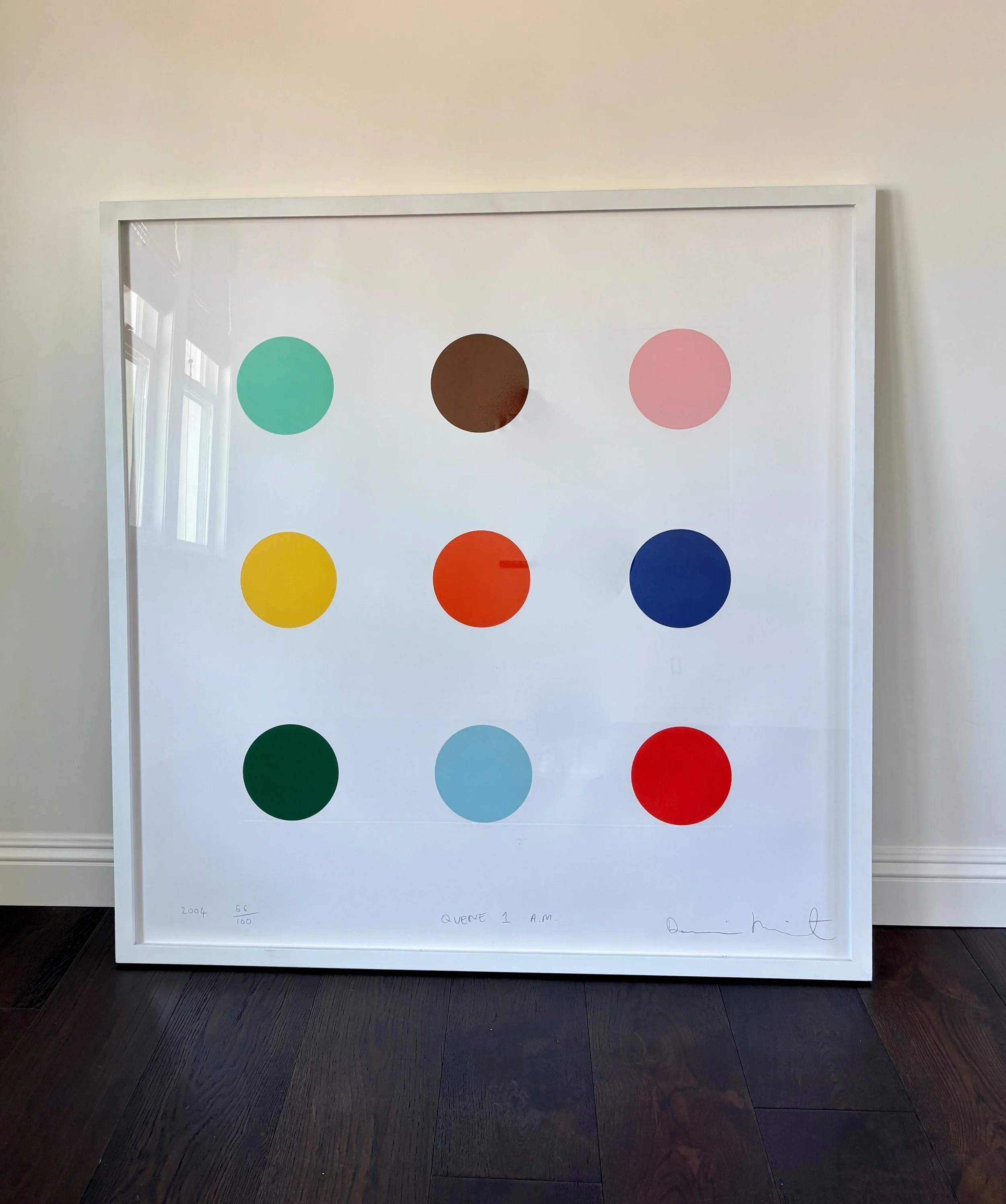 A framed artwork by Damien Hirst with a 3x3 grid of colored circles on a white background, placed on a dark hardwood floor against a light-colored wall.