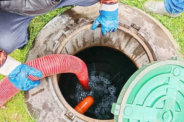 Two workers wearing gloves are inserting a red hose into an open manhole, with water flowing out.