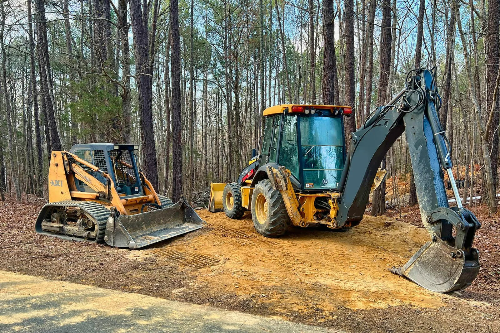 Construction equipment including a small bulldozer and an excavator parked in a wooded area with dirt ground.