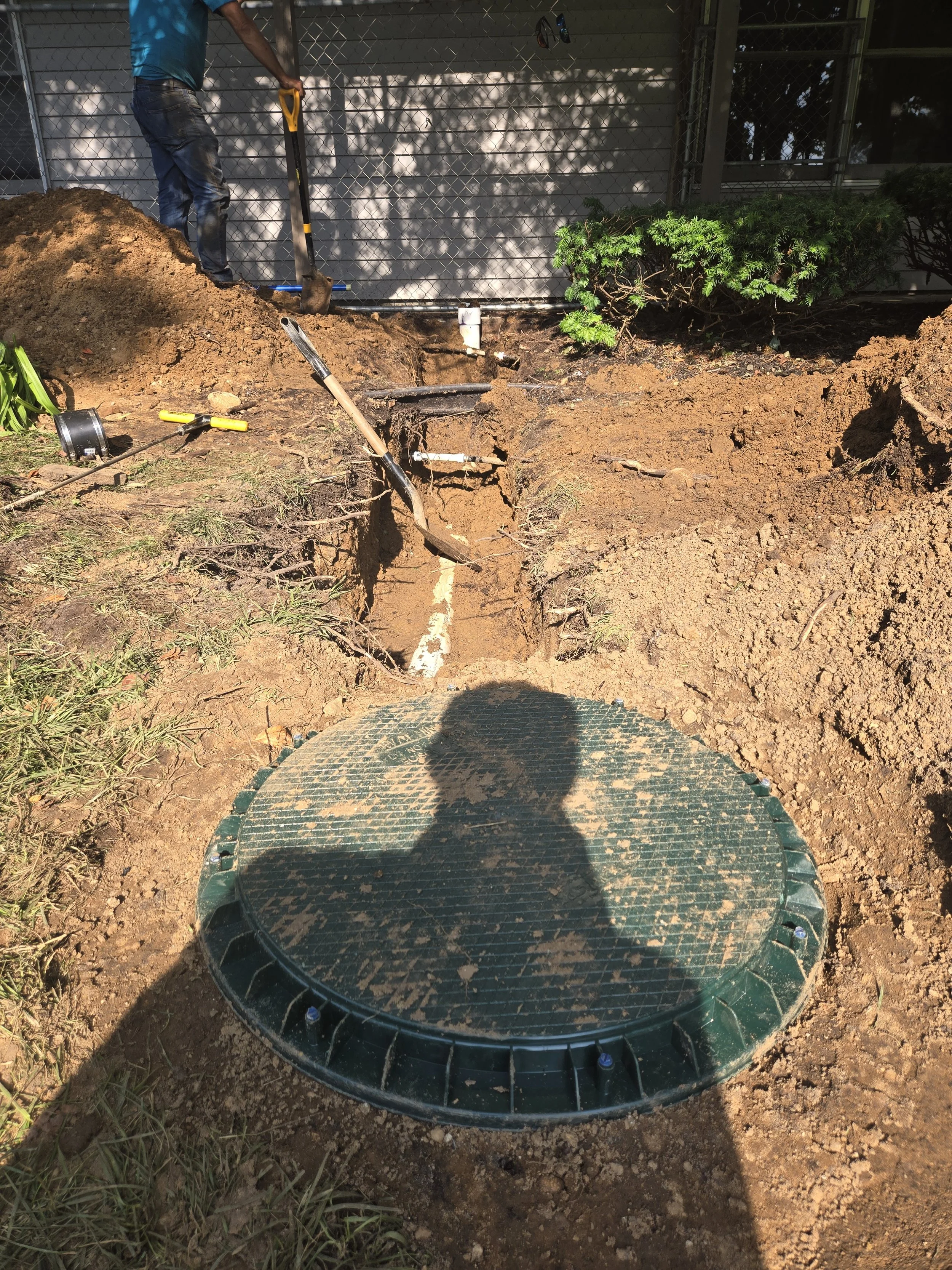A worker is installing or repairing a sewer or water line underground. The area around the manhole cover is excavated, with tools and pipes visible nearby.