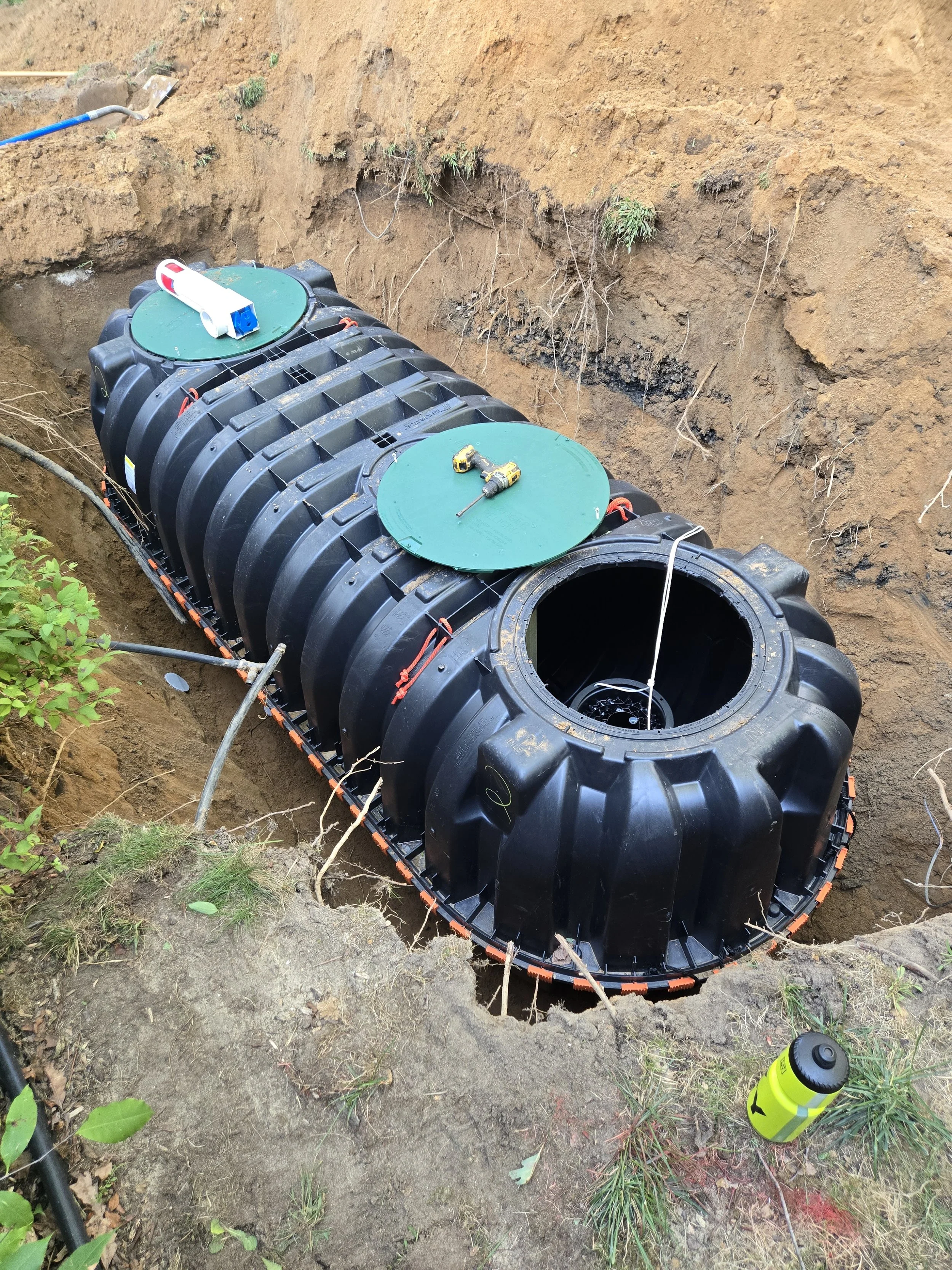 Underground black plastic septic tank in a excavation hole, with green access covers, tools, and equipment on top, at a construction site.