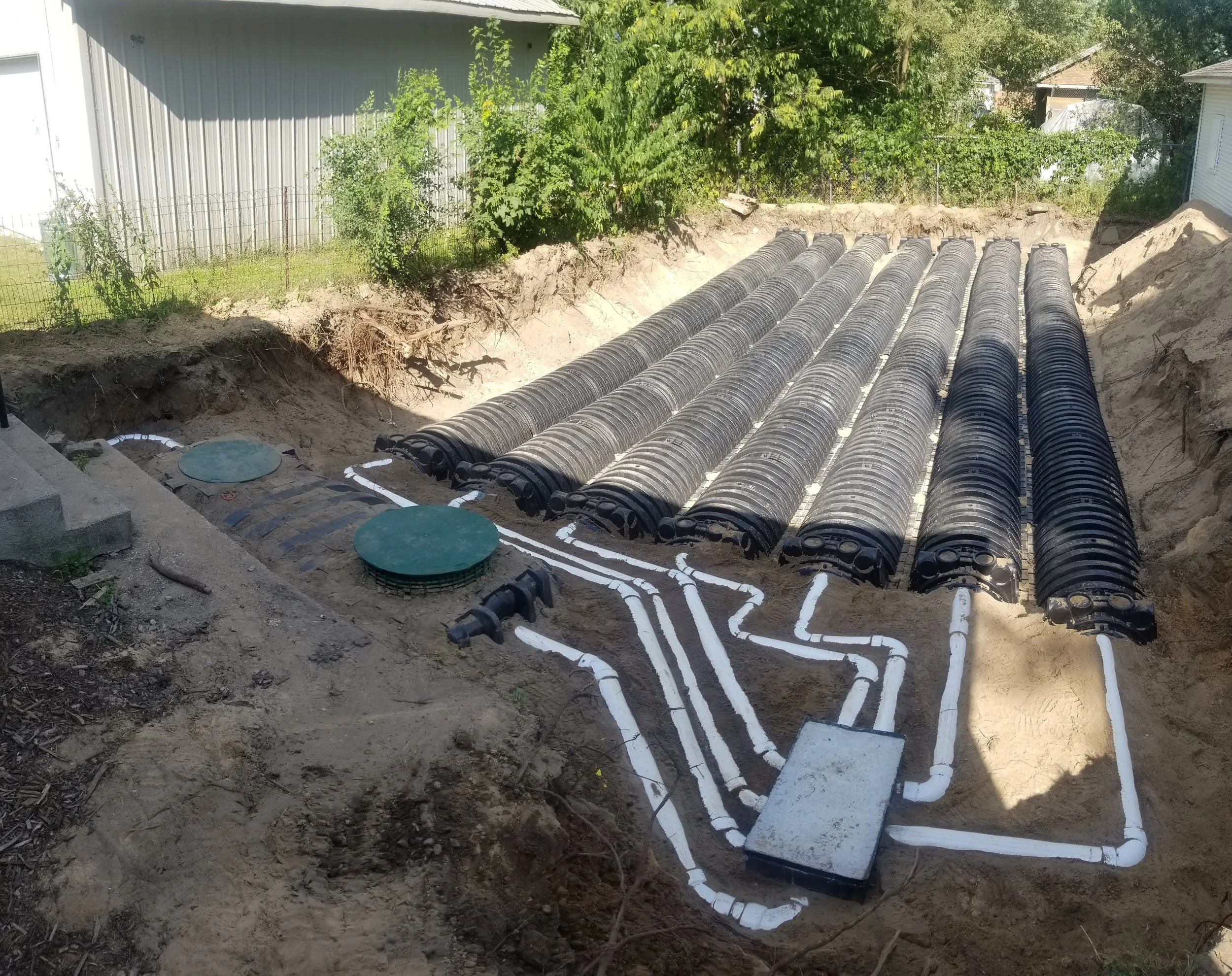 Underground piping for a construction project with black corrugated pipes and white PVC pipes laid in an excavated trench, with access covers and connection points visible.