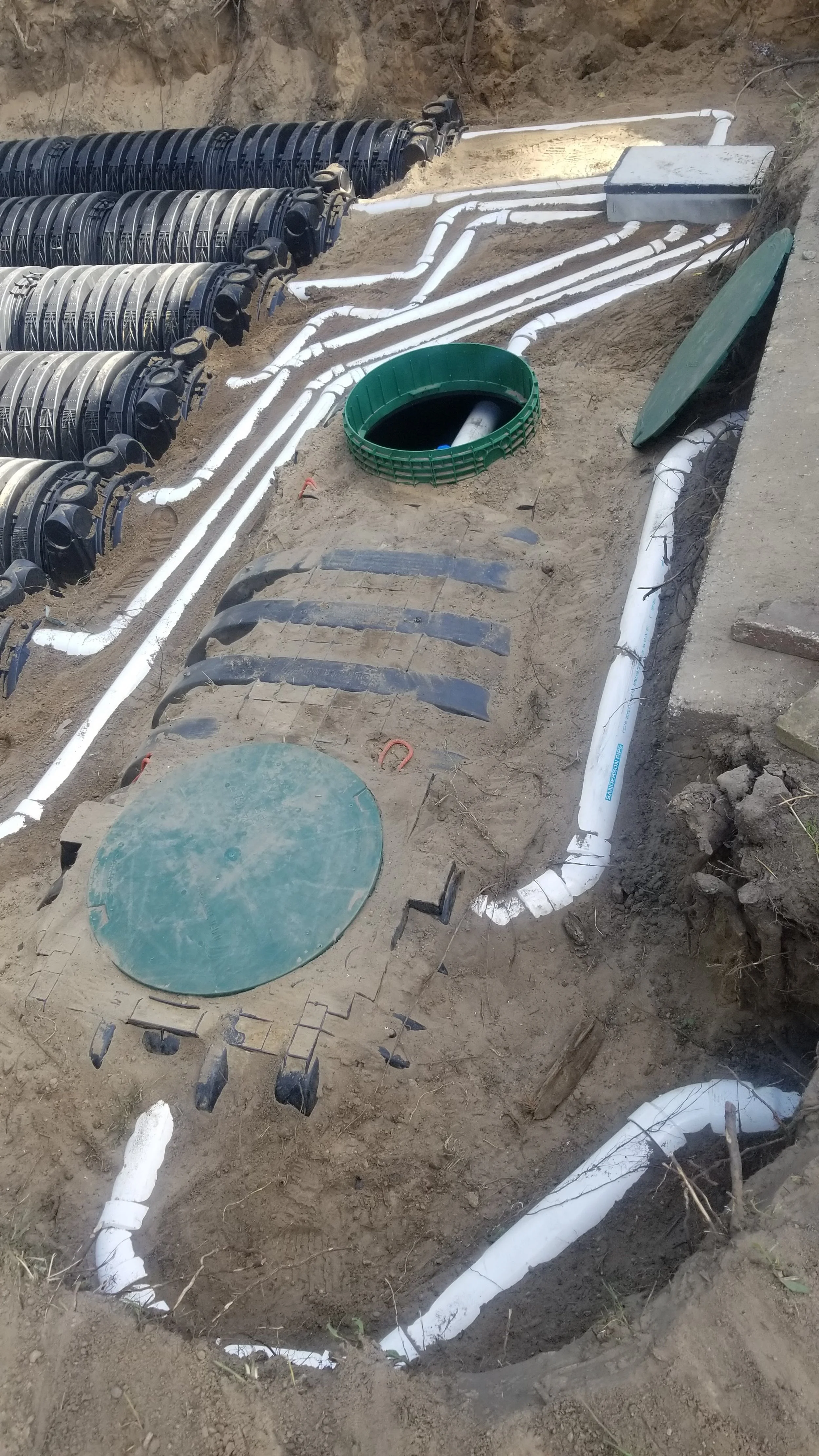 Underground plumbing and drainage pipes installation in soil, with black and white pipes, a green access cover, and flexible drainage fittings.