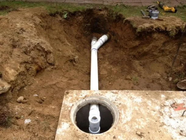 Underground plumbing pipe installation with a white PVC pipe set into a concrete pit, surrounded by dirt and construction tools.