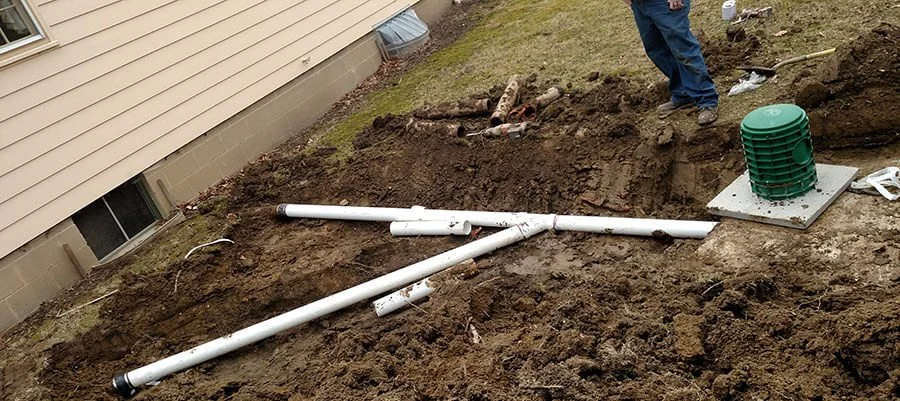 Underground water drainage pipes being installed outside a house, with a worker standing nearby and a green drainage component placed on a concrete base.