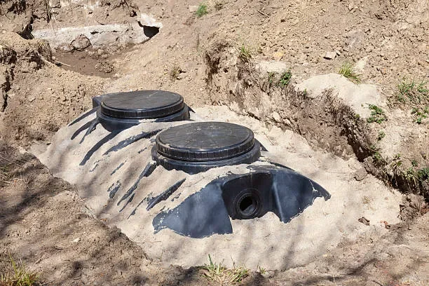 Two black septic tank lids partially buried in the ground, with a pipe connection nearby.