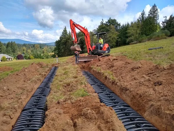 Excavator digging trenches for underground pipes in a grassy field with trees and mountains in the background.