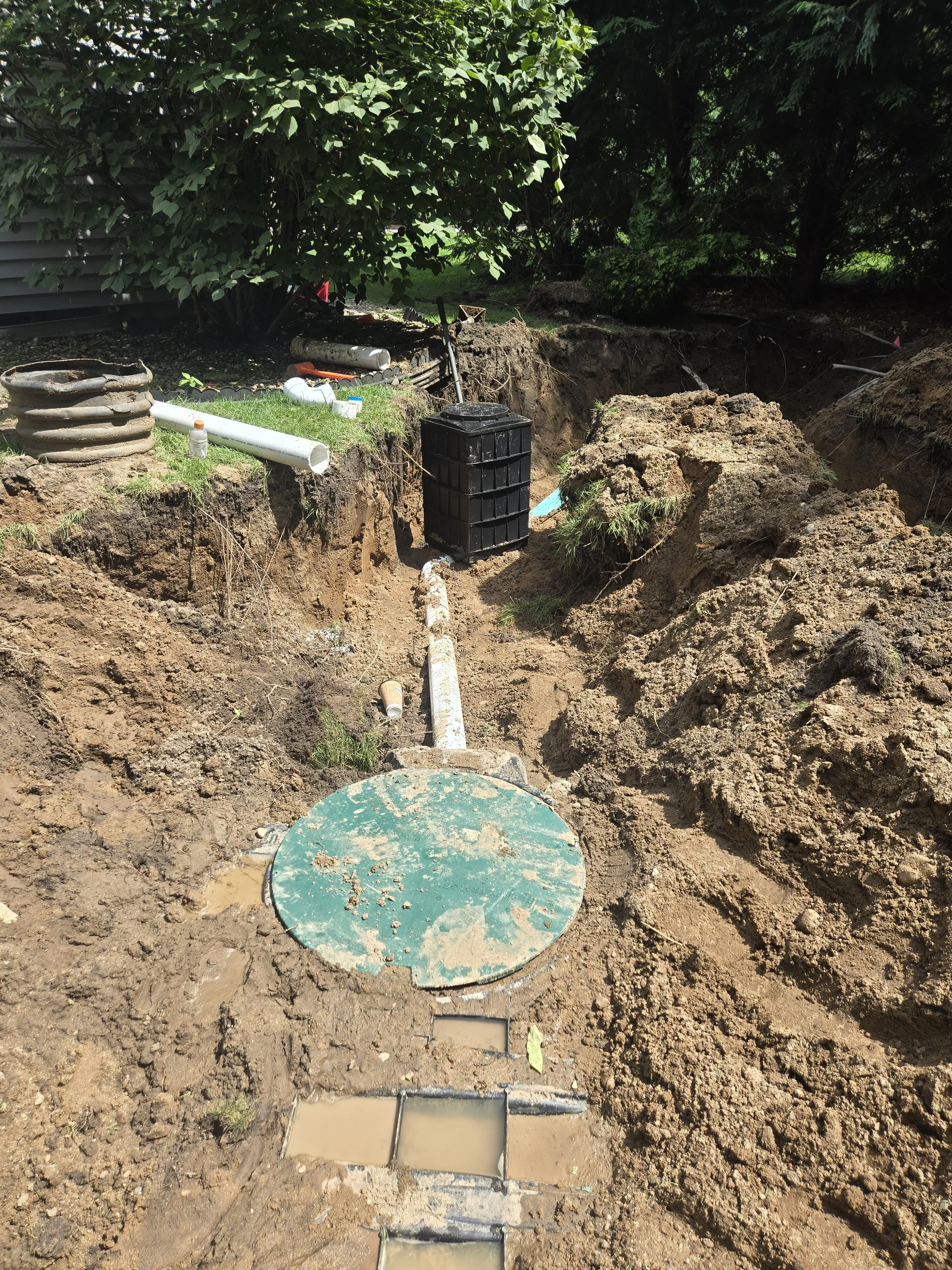 A construction site showing an underground plumbing installation with pipes, a black water collection tank, and a partially excavated trench.