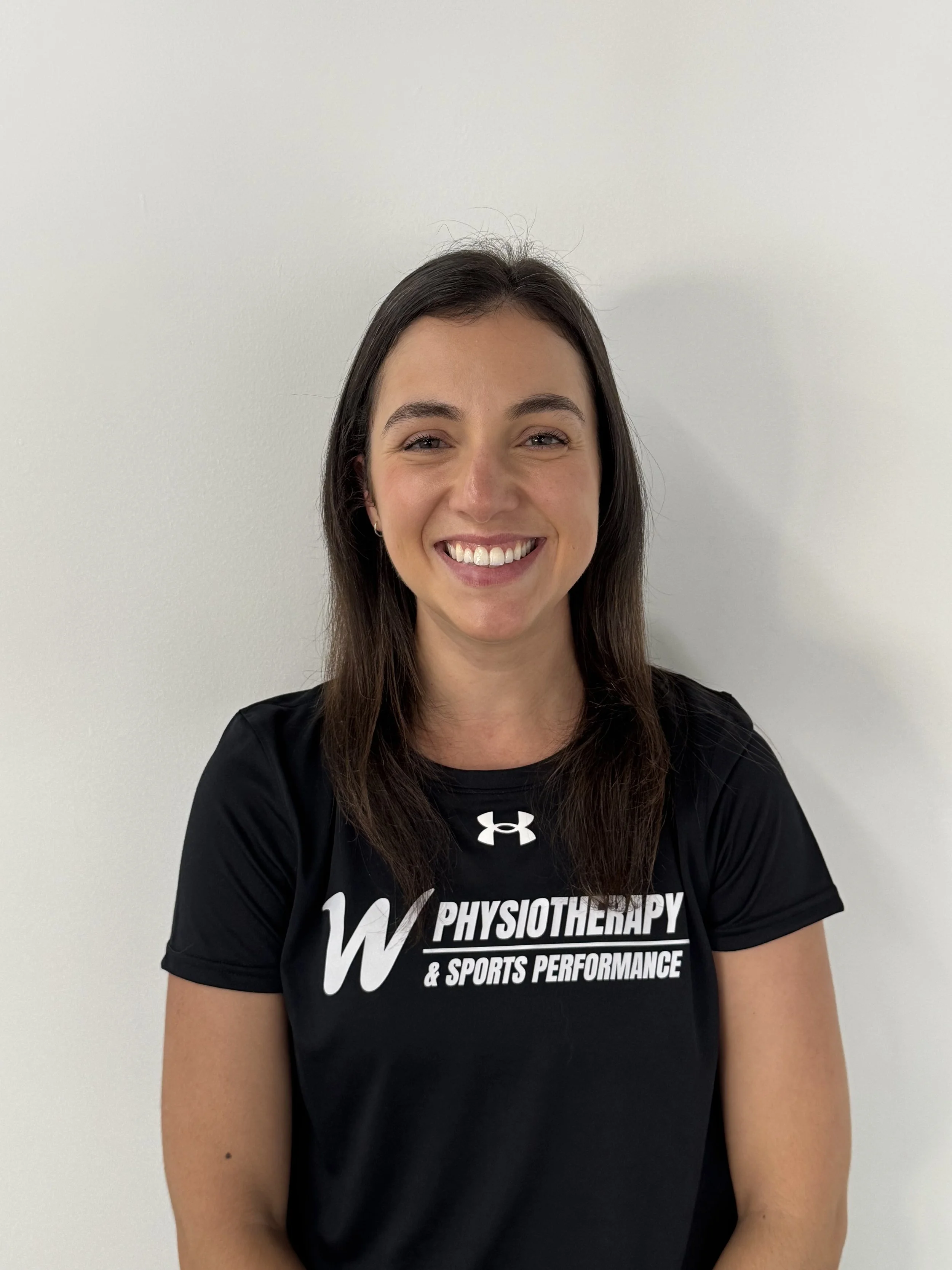 Smiling woman with long dark hair wearing a black Under Armour shirt with 'Physiotherapy & Sports Performance' written on it, standing against a white wall.